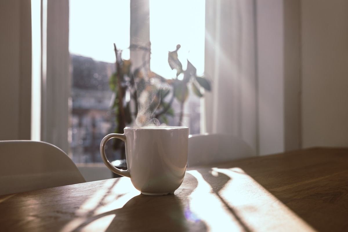 Taza humeante sobre una mesa de madera frente a una ventana iluminada por el sol