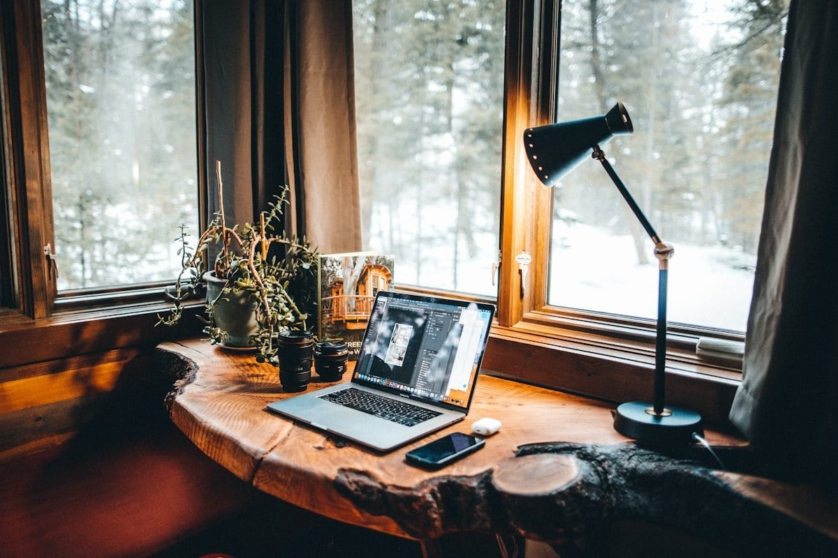 Portátil abierto sobre un escritorio de madera junto a una ventana con vistas a un bosque nevado