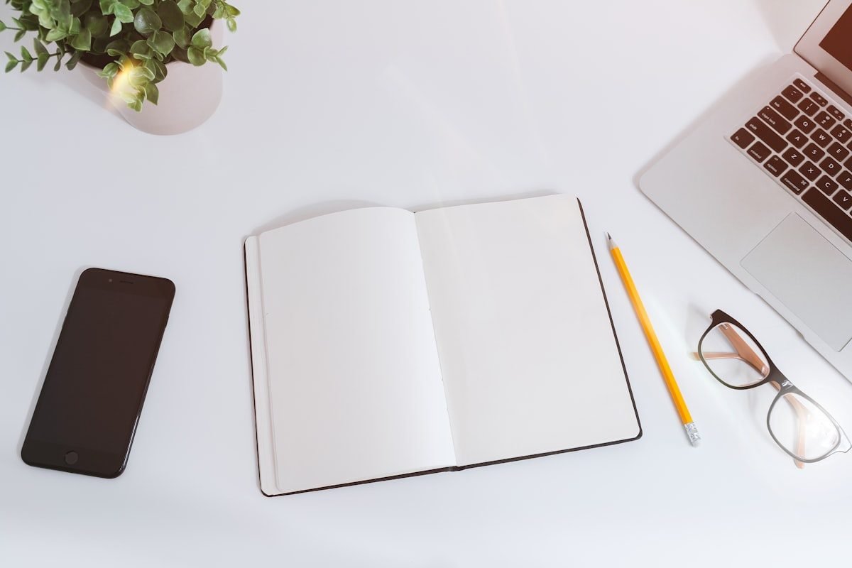 Cuaderno abierto en blanco sobre un escritorio junto a un portátil, un móvil y unas gafas